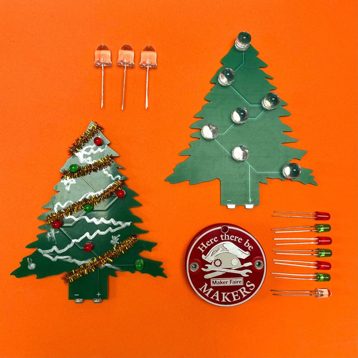 Learn To Solder - Holiday Tree soldering kit with green PCB trees, LEDs, and maker badge on orange background.
