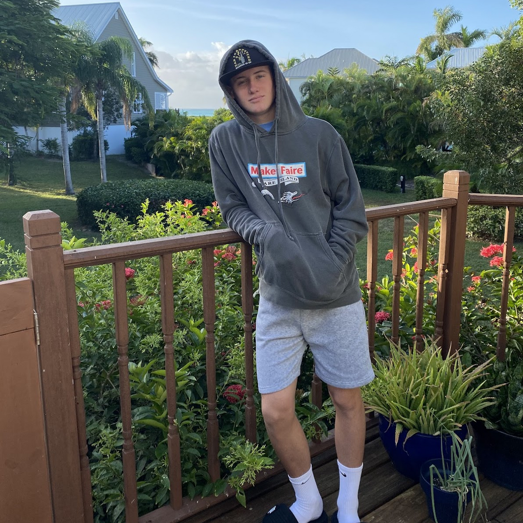 Person wearing a Maker Faire Mare Island hoodie and shorts on a deck with plants and a house in the background