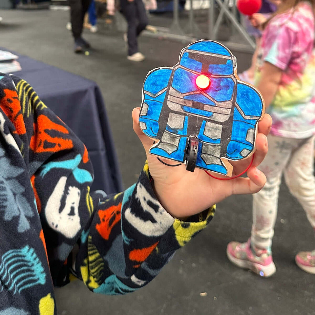 Child wearing a colorful shirt with a cartoon design, holding a blue robot shaped craft with a red LED