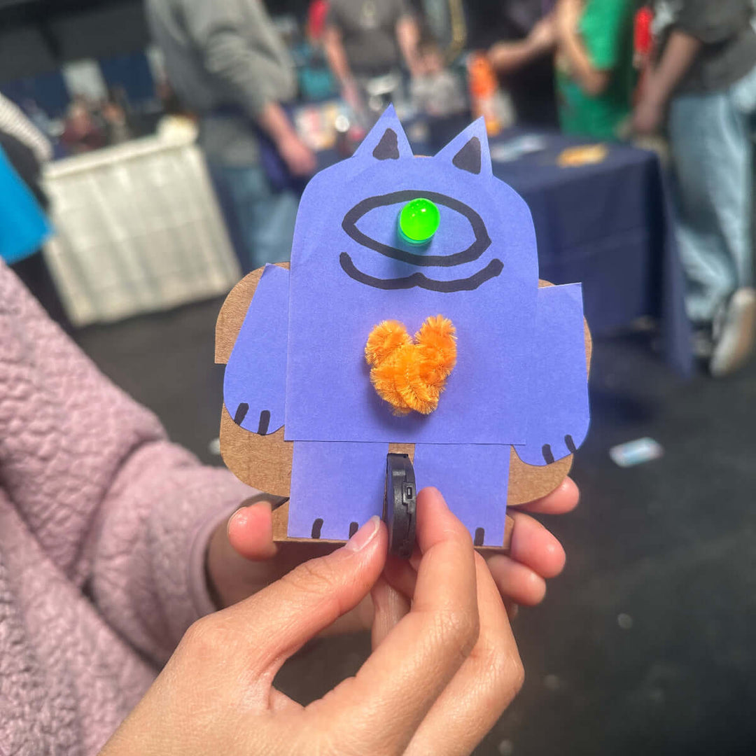 Hands holding a small blue cat-shaped craft project with orange tufts for a heart and a single LED eye