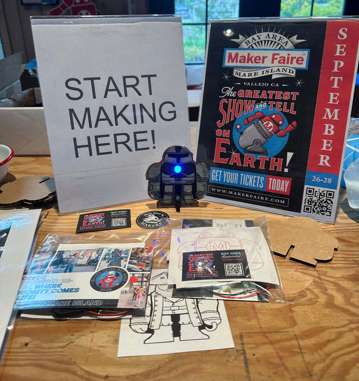Tabletop display with Maker Faire promotional materials and a small robot-like craft project with an blue LED light.