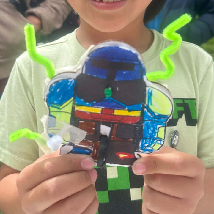 Child holding a colorful craft project in front of a Maker Faire tent outdoors