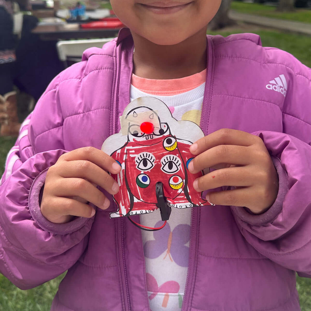 Child in a purple jacket holding a cardboard  project outdoors at Maker Faire