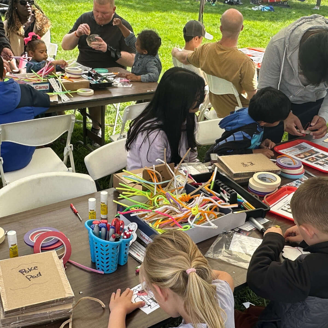 Children and adults engaged in craft activities outdoors on a sunny day at a Maker Faire event.