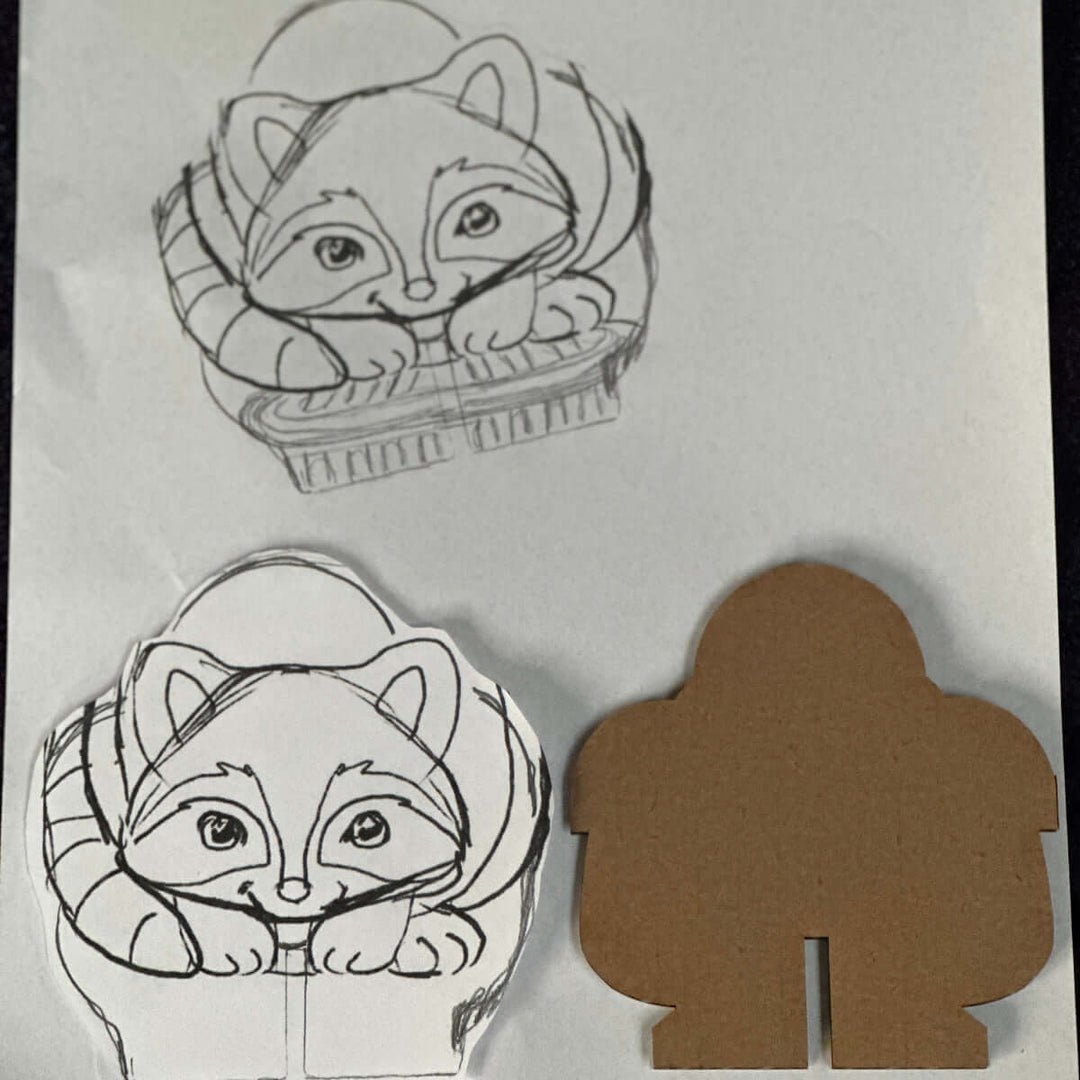 Original cartoon raccoon design on a white card next to a brown cardboard cutout.