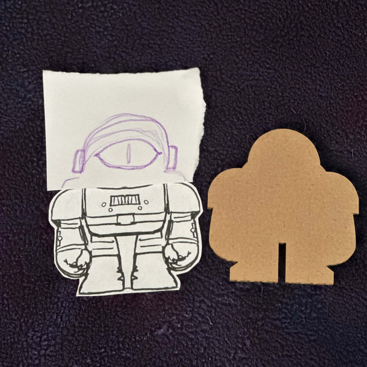 Cardboard cutout next to a hand drawn one eyed robot astronaut on a black background