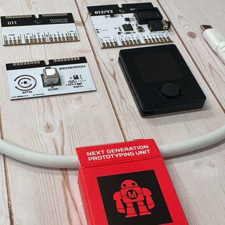 Oxocard Connect 2 kit laid out on a light wood surface, including a red retail box with Makey the robot, and three white and black boards with a USB-A to USB-C white cable.