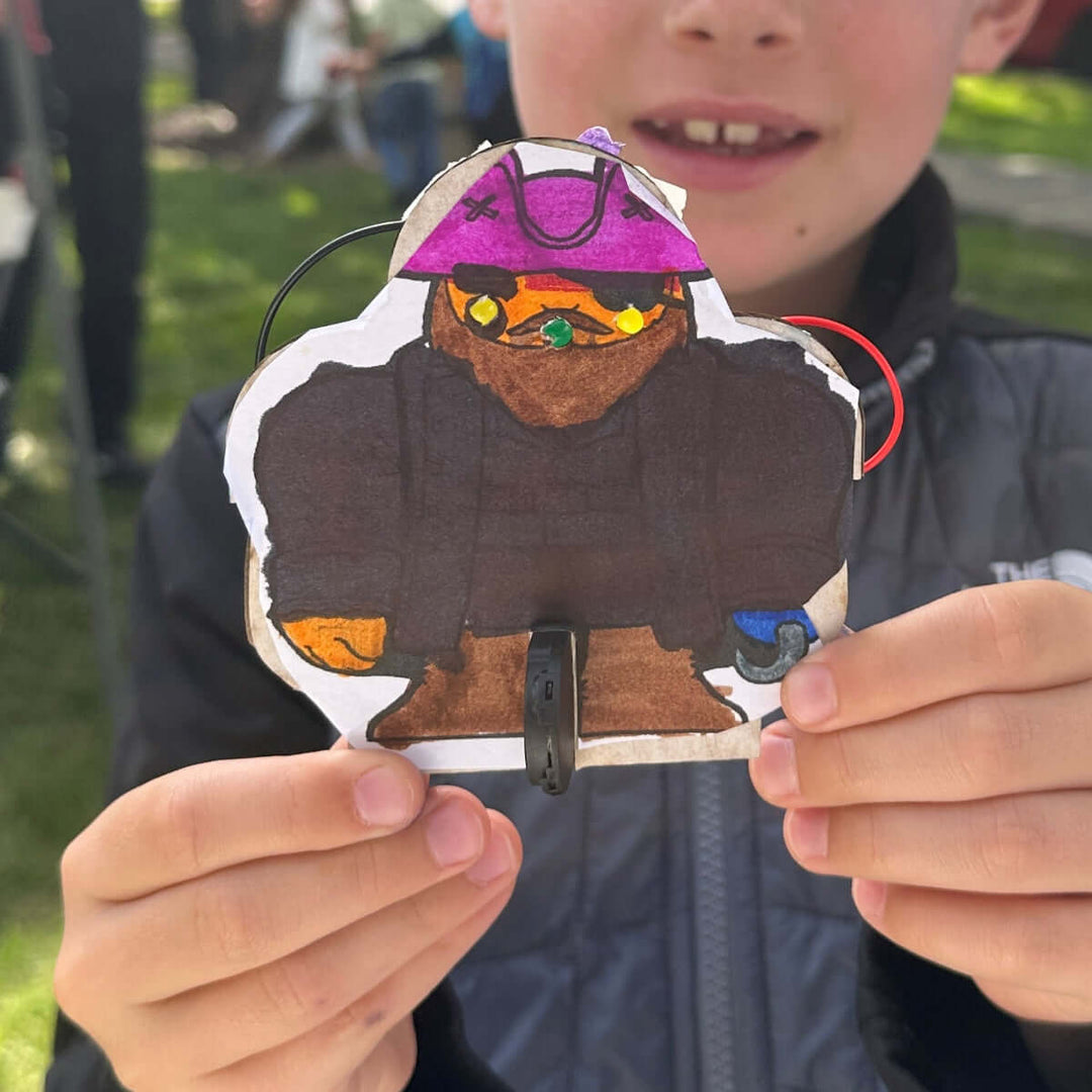 Child holding a small pirate cardboard LED project outdoors at a Maker Faire event with trees and grass in the background